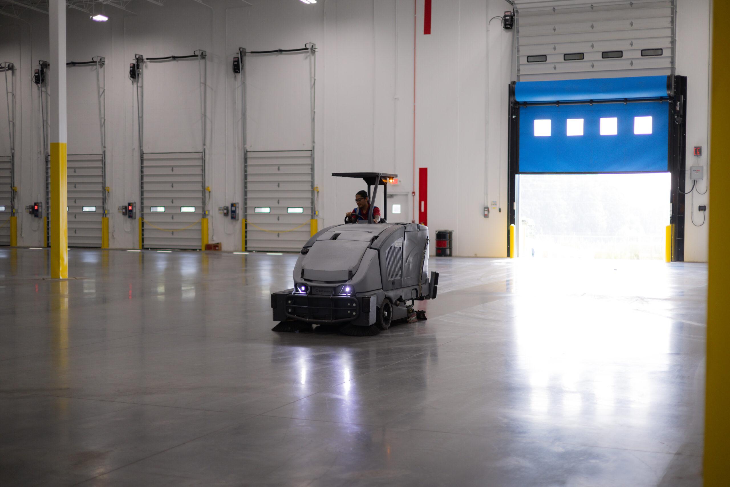 MFS technician operating a ride-on floor scrubber inside a large warehouse facility